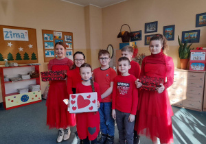 Pięcioro młodszych uczniów stoi pośrodku sali, są ubrani na czerwono. Po bokach stoją dziewczynki ze starszej klasy i trzymaja skrzynki na kartki walentynkowe.